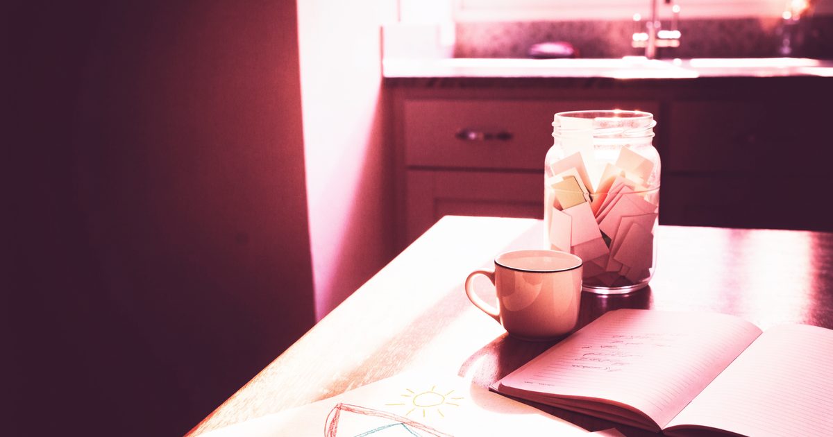A sunlit kitchen table with an open notebook, a coffee mug and a glass jar filled with small folded paper notes, in soft pink morning light.